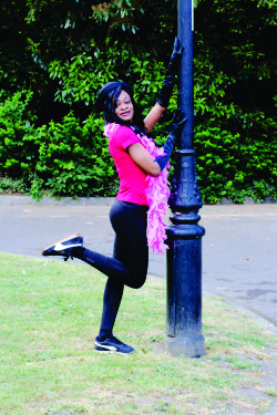 Elle in a red top, black leggings and a pink feather boa poses playfully as a Scandalous Character next to a lamppost outdoors.