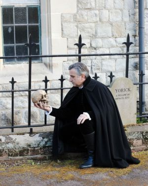 Mark in a black cape and costume kneels by a tombstone, holding a skull. The tombstone is engraved with YORICK A FELLOW OF INFINITE JEST.
